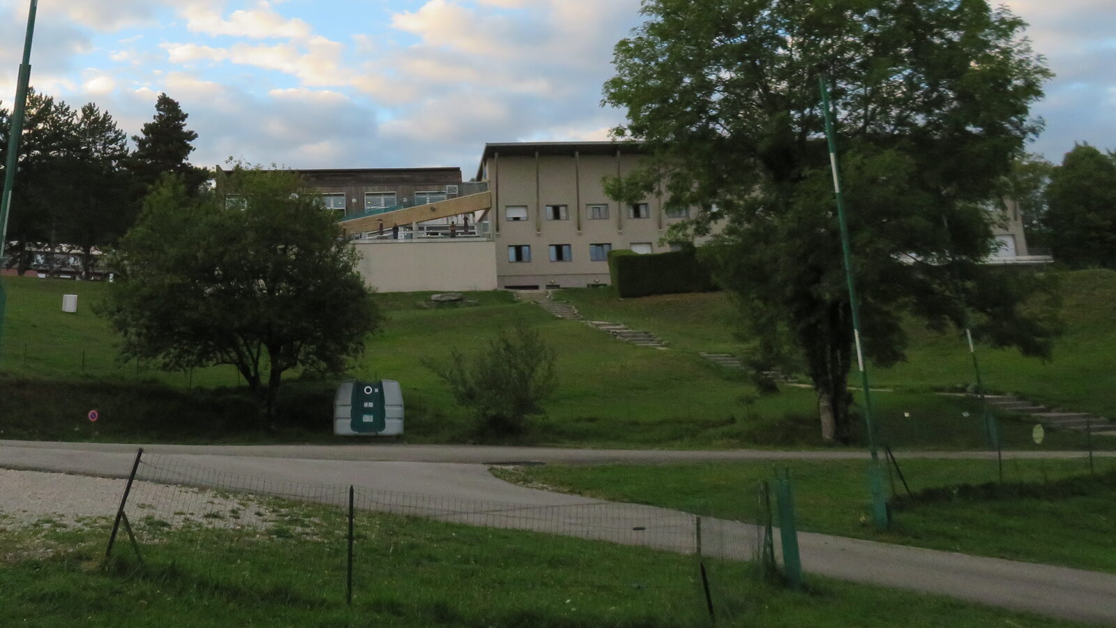 Les bâtiments principaux du centre sportif de Bellecin, longue façade claire à deux niveaux en haut d'une prairie en pente bordée d'arbres.