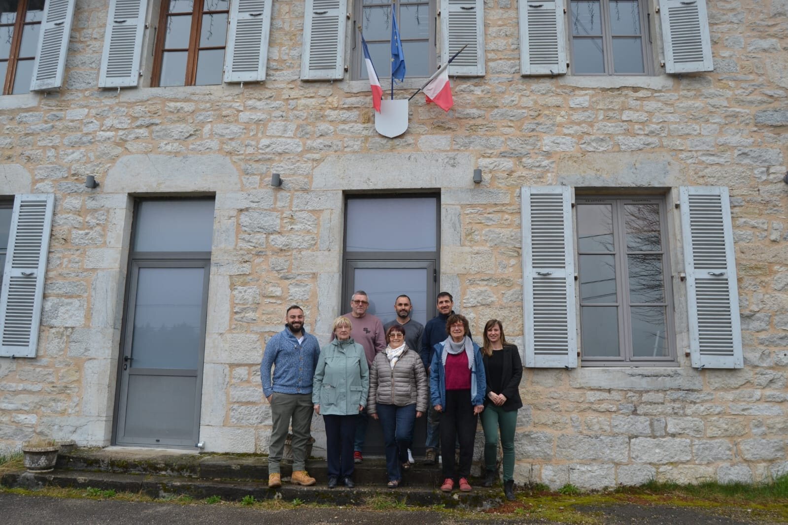 Les sept conseillers municipaux de Vertamboz alignés devant le perron de la mairie, façade en pierre claire, drapeaux français et européen au-dessus de la porte.