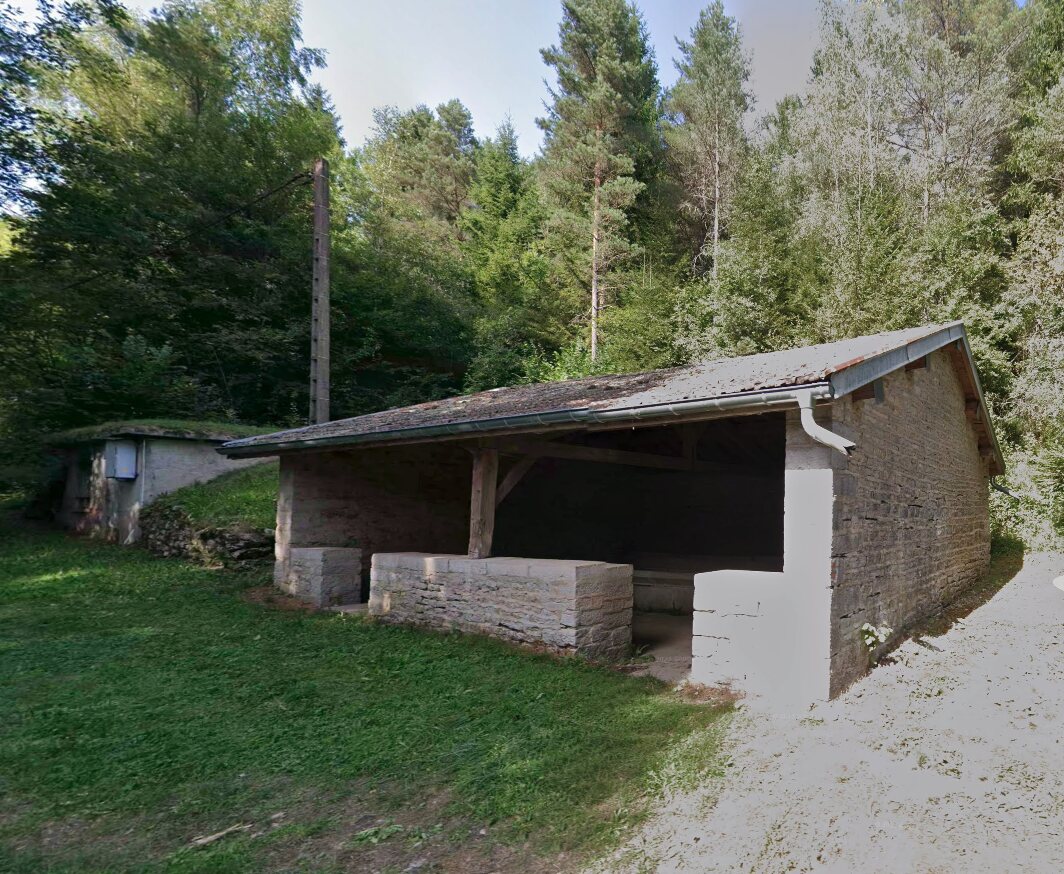 Le lavoir de Vertamboz : bâtiment en pierre claire avec toit en tuiles, abrité sous une lisière de sapins, bassins en pierre visibles devant.