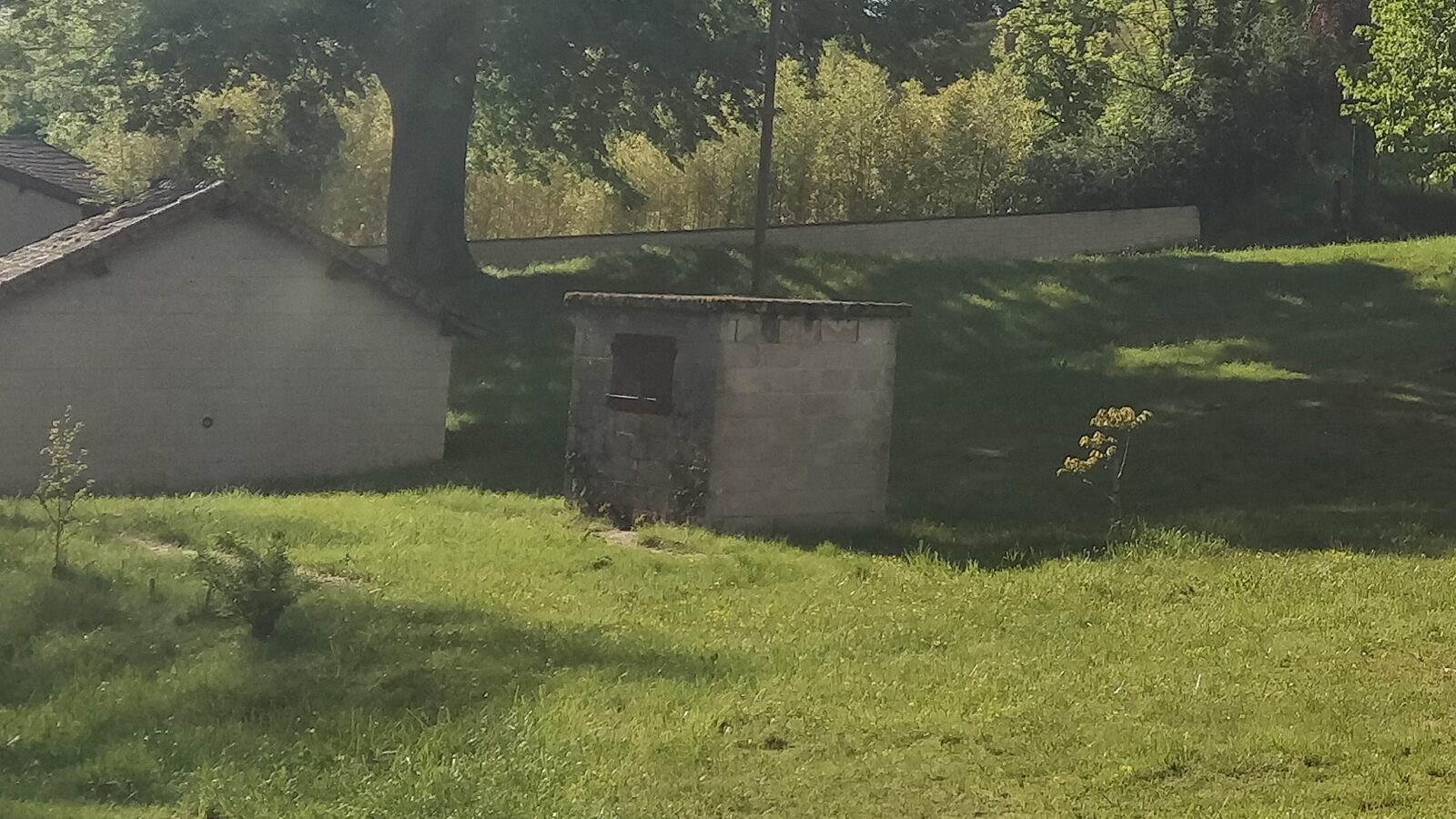 Le poids public de Vertamboz : petite construction en pierre à toit plat et porte de fer, au bord du pré où se tiendra la fête, à côté d'un hangar communal et sous un grand arbre.