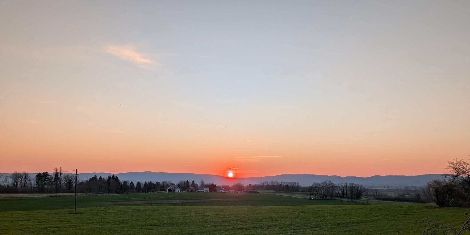 Coucher de soleil orangé sur les crêtes du Jura, prairie en pente au premier plan, lieu-dit « Sous la Côte ».